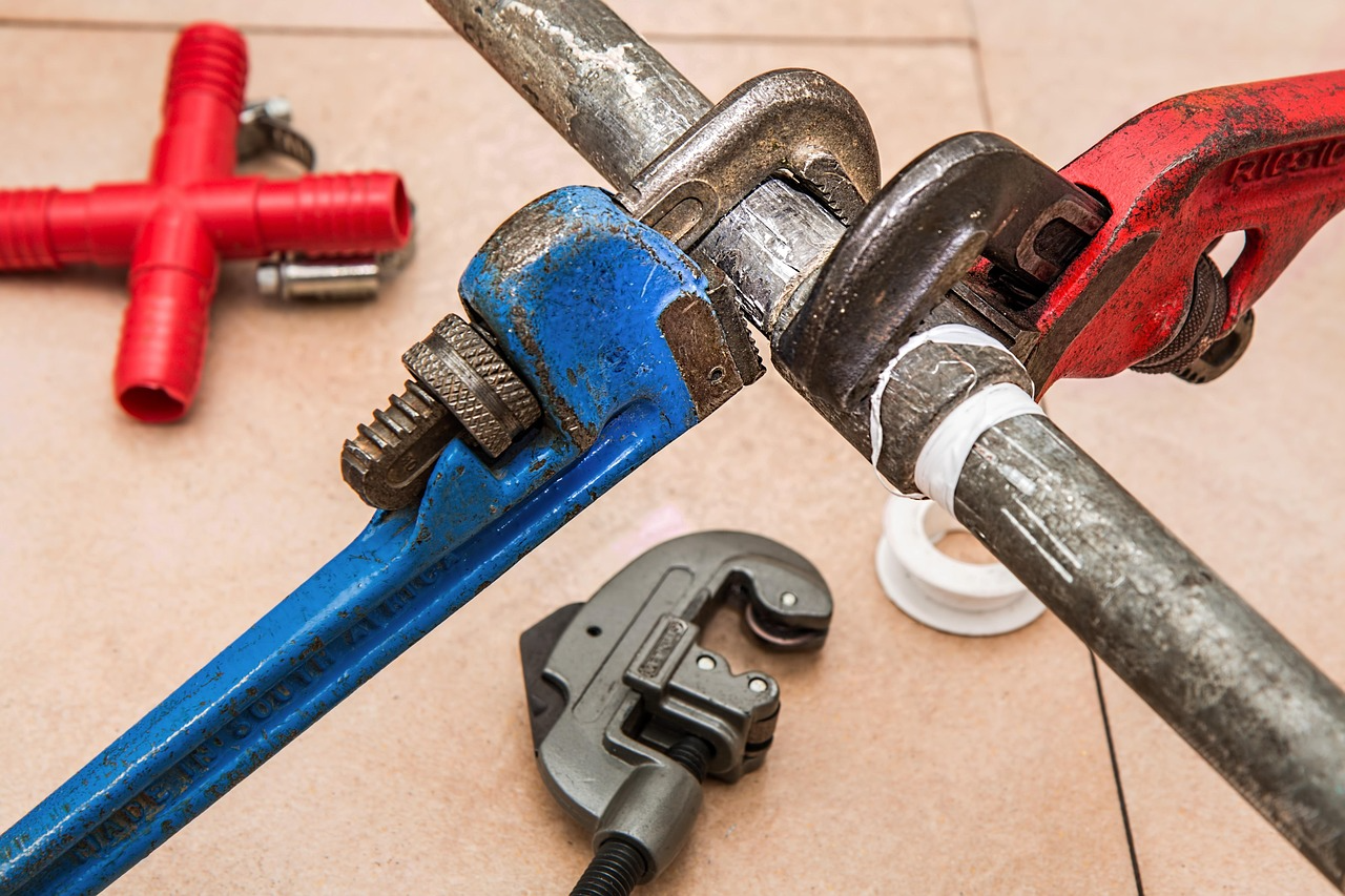 Plumbing tools placed on the floor beside a metal pipe during repair work.